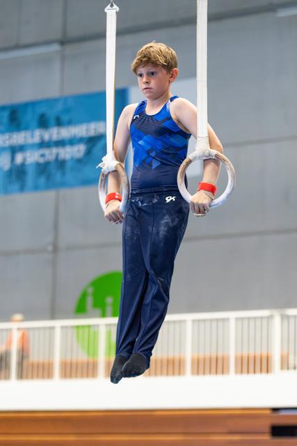 Young athlete performing on still rings with focused expression, wearing blue geometric-patterned leotard at indoor gymnastics facility