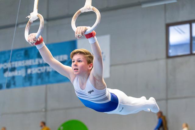 Young athlete performs on rings with extended body position, wearing white and blue outfit in indoor training facility