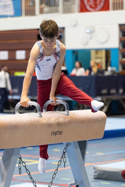 Young athlete performing on pommel horse with intense concentration, wearing red and white uniform at indoor gymnastics meet