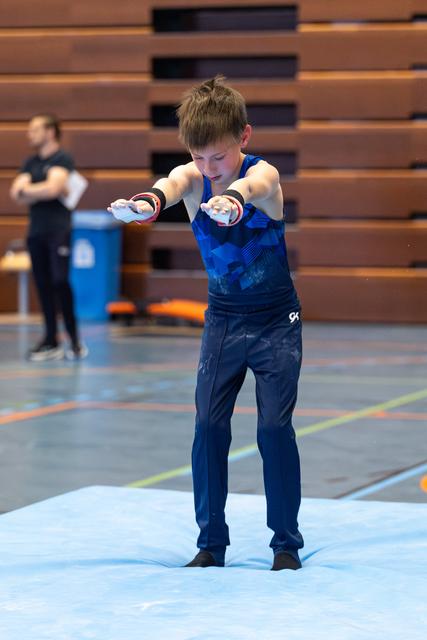 Young athlete in blue tank top practices balance with arms extended forward, standing on light blue mat in gym
