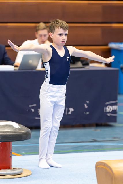 Young athlete in navy leotard and white pants prepares with arms extended on competition floor