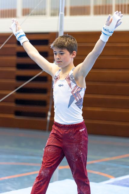 Young gymnast in white and red uniform raises arms in victory pose on competition floor, displaying focused determination