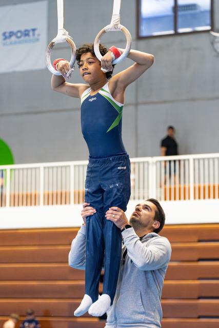 Young athlete performs still rings routine while coach spots from below in indoor training facility