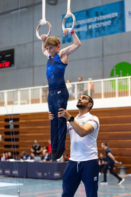 Young athlete performs on rings while coach spots from below during indoor training session
