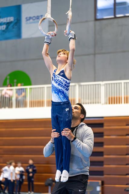 Young athlete performs rings exercise with coach supporting below in indoor gymnasium during training session