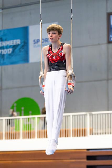 Young athlete hanging on still rings during routine, displaying focused concentration in white uniform at indoor venue