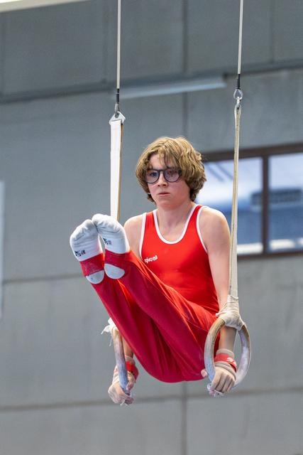 Young athlete with glasses performs on rings, displaying focused determination in red uniform during indoor event