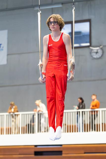 Young athlete in red uniform and glasses performing a still rings hold during indoor training session