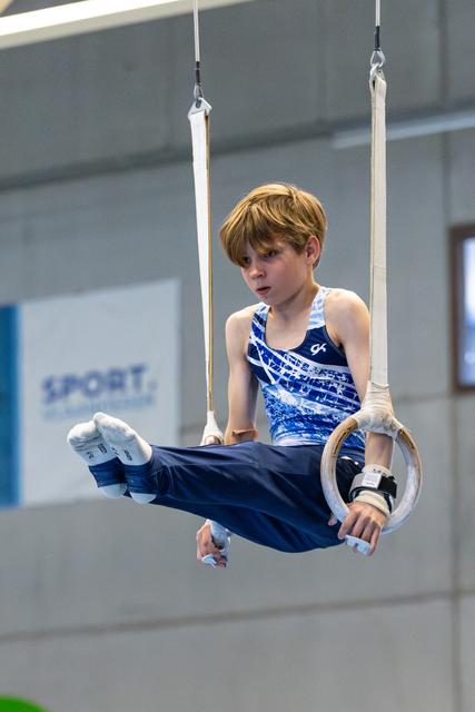 Young gymnast performing on still rings with focused concentration during training, wearing blue patterned leotard