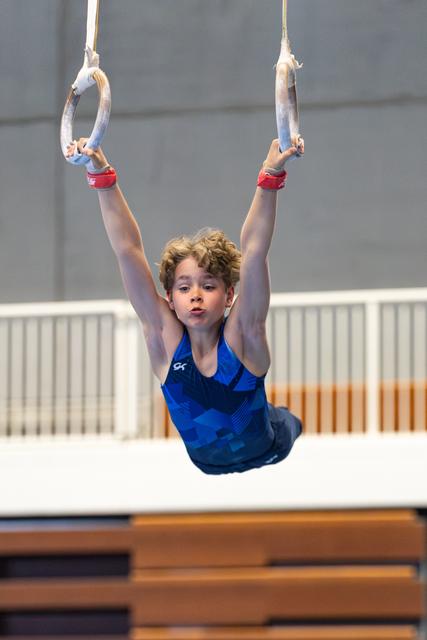 Young athlete performs a hang on still rings with extended arms, wearing red grips and blue leotard, focused expression