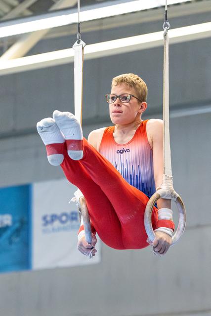 Young athlete performing on still rings with focused expression, wearing glasses and an orange-to-blue gradient leotard