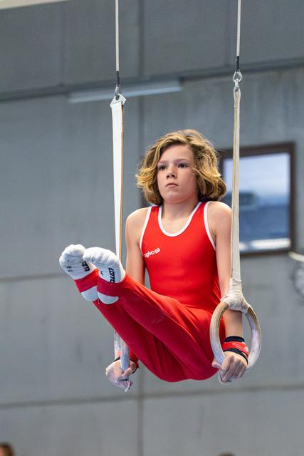 Young gymnast performing on rings with intense concentration, wearing red uniform and hand grips in training facility