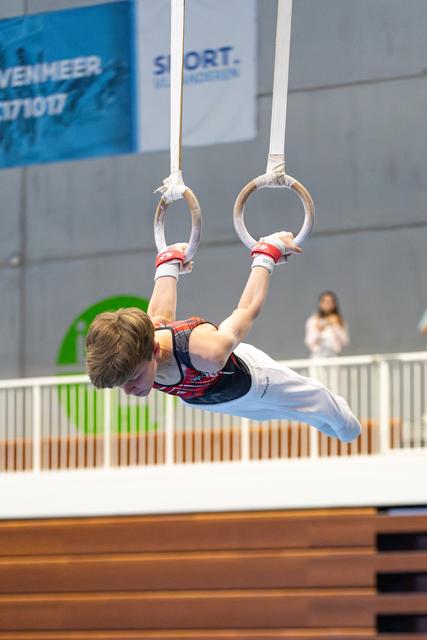Young athlete performing an inverted hang on still rings during indoor competition, demonstrating upper body strength