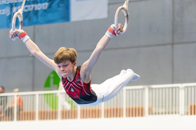 Young athlete performs rings routine with arms extended overhead, demonstrating strength and concentration in competition setting