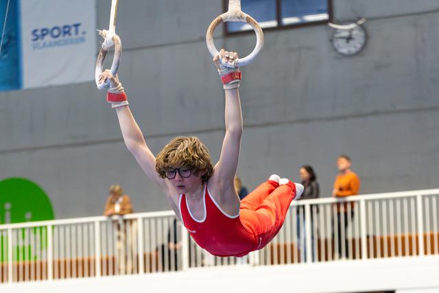 Young athlete in red leotard performing on still rings with protective grips, wearing glasses during indoor training