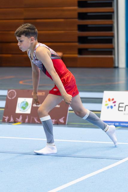 Young athlete in red shorts running across blue floor mat during routine, focused expression, wooden bleachers in background