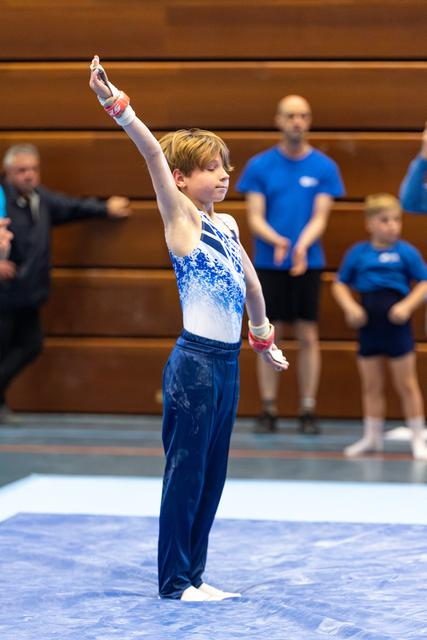 Young gymnast in blue and white leotard strikes a confident salute pose during floor exercise with spectators in background
