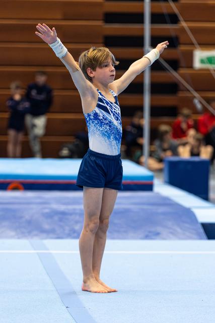 Young gymnast in blue and white leotard stands with arms raised in finishing pose on floor exercise mat