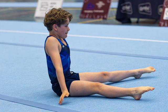 Young athlete in blue leotard performs seated pike stretch on mat, legs extended straight with focused concentration