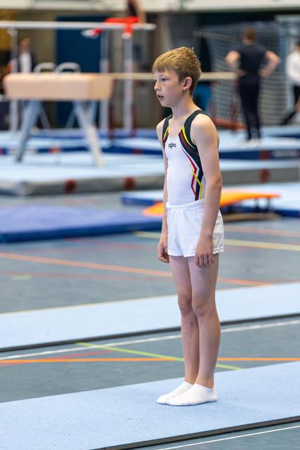 Young gymnast in white leotard stands at attention on the floor, focused and ready before his routine