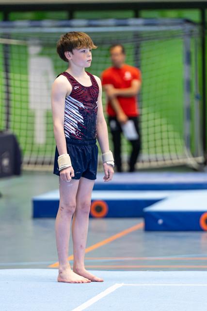 Young gymnast stands at attention on floor mat, focused expression, wearing sequined leotard with wrist supports
