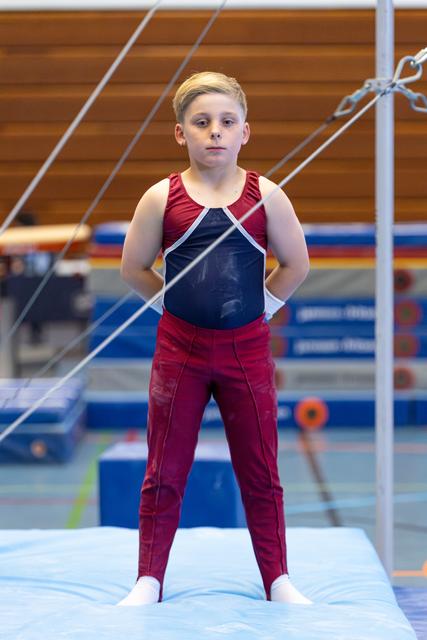 Young athlete in maroon and navy leotard stands with focused expression on blue mat, ropes visible in foreground