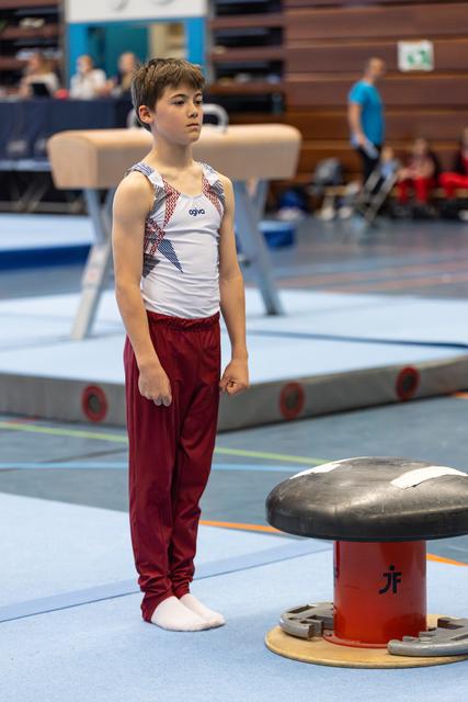 Young gymnast in white and red uniform stands attentively beside pommel horse, preparing for his routine in competition hall