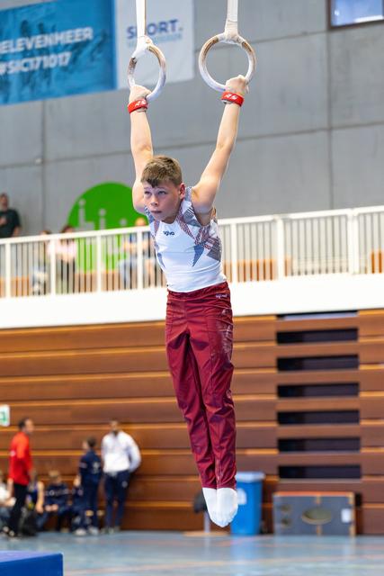 Young athlete hangs from still rings with focused expression, wearing white and maroon uniform during indoor competition
