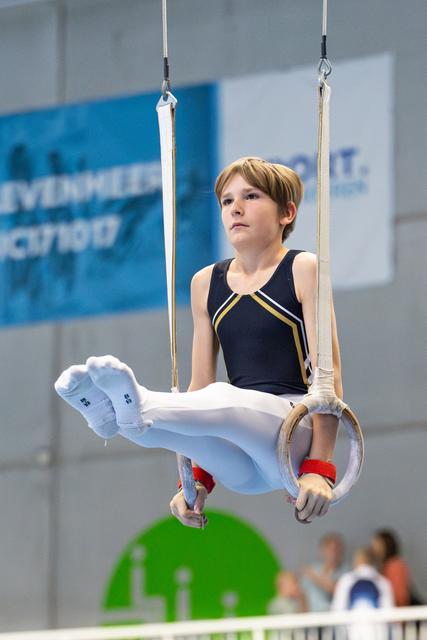 Young athlete performing on still rings with focused expression, wearing navy leotard with white pants and red wristbands