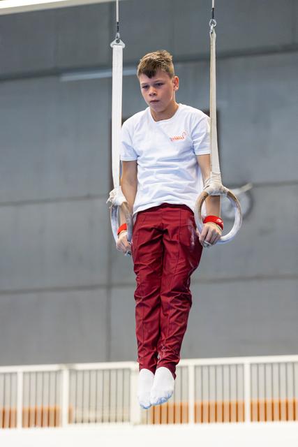 Young athlete hanging from still rings in white shirt and red pants, showing intense focus during training or performance