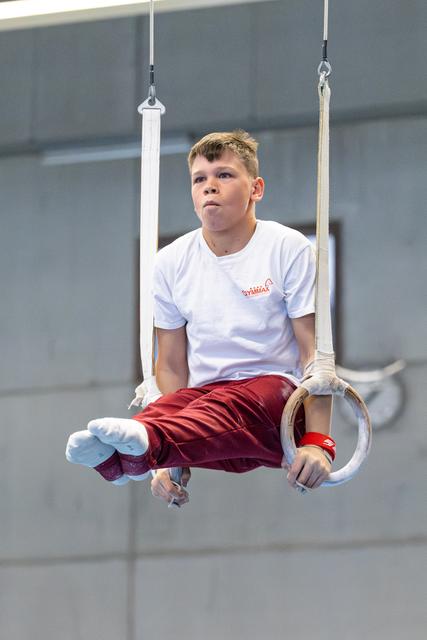 Athlete performing still rings hold, focused expression, white shirt and burgundy pants, indoor training facility