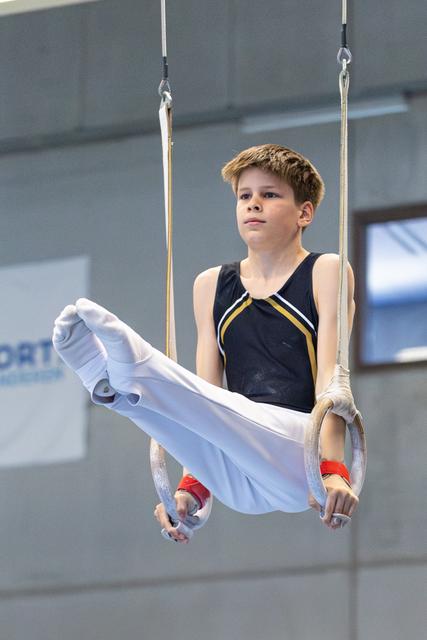 Athlete performing still rings routine with extended legs, wearing black and yellow leotard in training facility