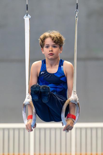 Young athlete performing a still rings routine with intense focus, wearing blue uniform and white grips during competition