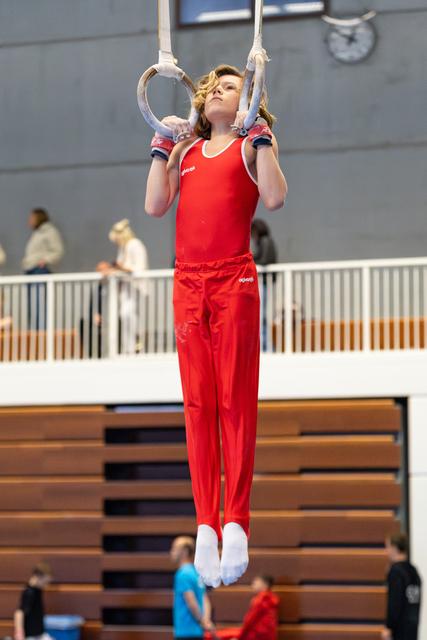 Young gymnast performs on still rings in red uniform, displaying focus and strength during routine at indoor sports facility