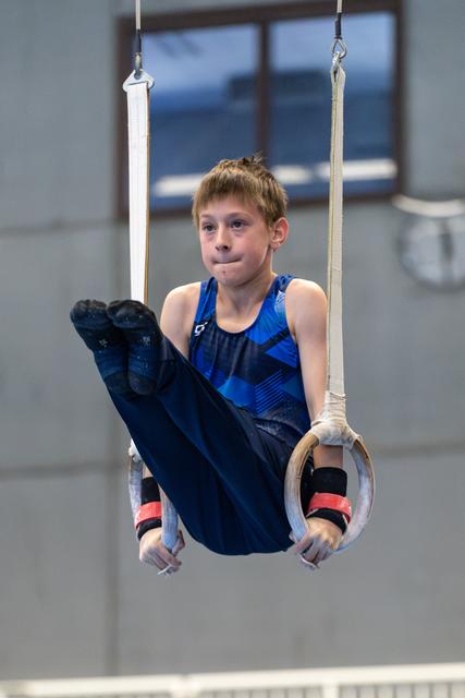 Young gymnast performing a L-sit hold on rings, legs extended forward, displaying concentration and strength in training facility