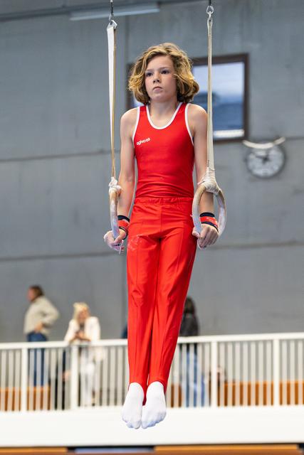Young gymnast in red uniform performs a hold position on still rings during competition at indoor venue