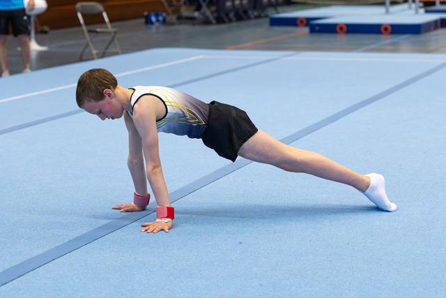 Young athlete in yellow and black leotard performs floor stretching exercise on blue mat, demonstrating flexibility and form