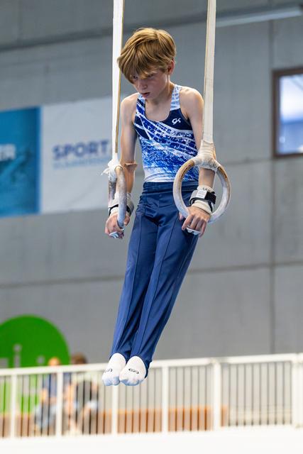 Young gymnast suspended on rings apparatus in blue gradient leotard, focused expression during training or competition