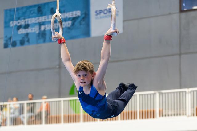 Young athlete swings on still rings with arms extended overhead, legs in horizontal position during training at indoor sports facility