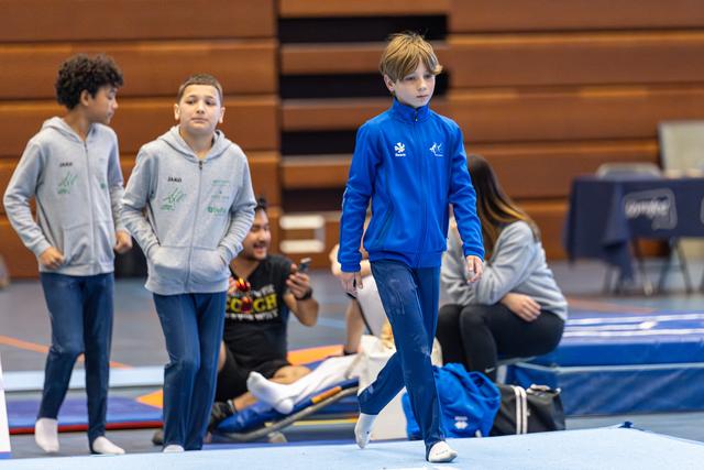 Young athlete in blue tracksuit walks across gym floor while teammates in gray warm-ups watch from the side