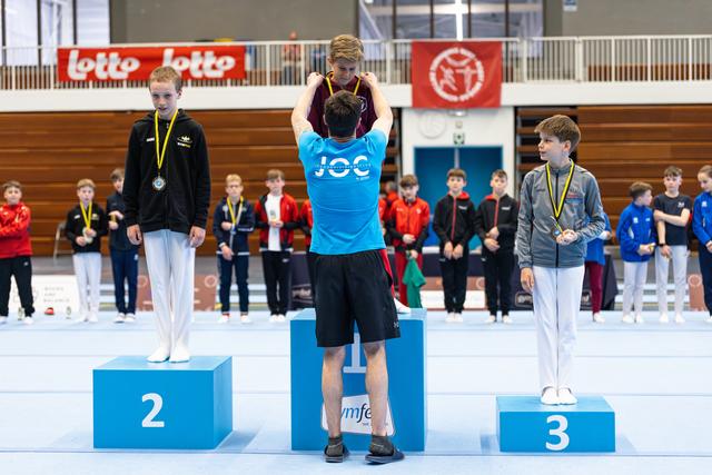 Three young athletes stand on award podium wearing medals, with other participants watching in the background at indoor sports hall