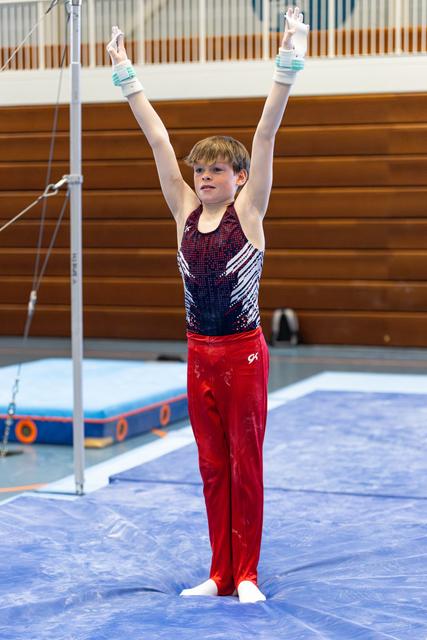 Young gymnast stands with arms raised in triumphant pose on mat, wearing sequined leotard and red pants in training facility
