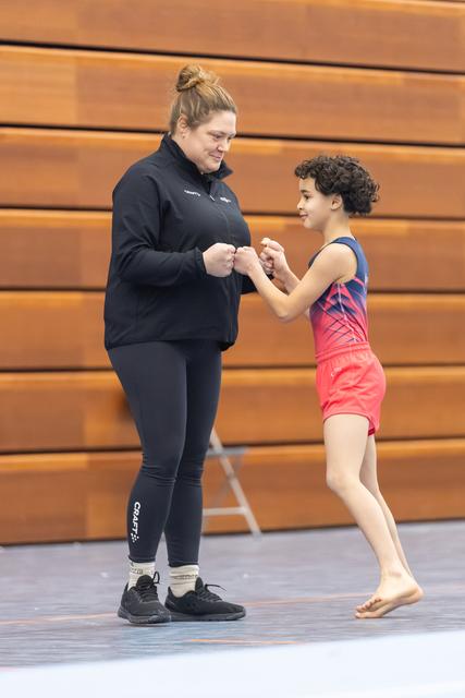 Coach in black sportswear shares a warm fist bump with young gymnast in pink leotard, both smiling in gymnasium