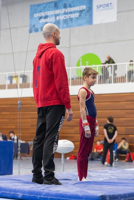 Coach in red hoodie stands beside young gymnast in maroon uniform on blue floor mat, observing the arena together