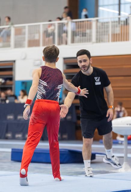Coach in black gestures toward a gymnast wearing a sparkly leotard and red pants on the competition floor