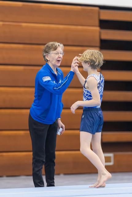 Coach in blue jacket offers guidance and encouragement to young gymnast in blue leotard during floor exercise training