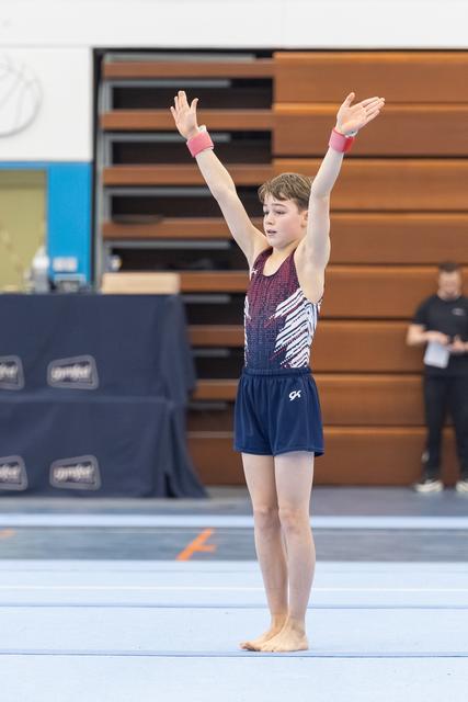 Young gymnast stands with both arms raised overhead on floor mat, performing routine in gradient leotard at indoor venue
