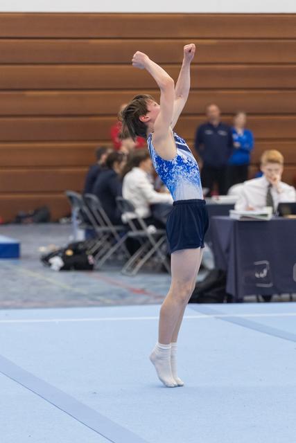 Gymnast stands on tiptoes with both fists raised triumphantly after completing floor routine, judges table visible behind