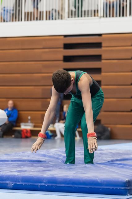 Male gymnast in green unitard bends forward on blue mat during floor routine, showing focused concentration in gym setting