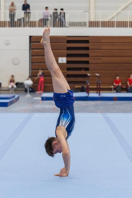 Young gymnast performs a handstand on floor mat, legs extended upward, with spectators watching from bleachers